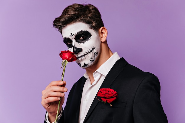 Homme brune en smoking posant avec rose à l'halloween. Beau modèle masculin avec maquillage effrayant mexicain debout sur fond violet.