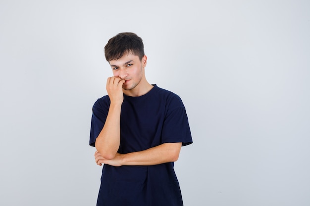 Homme brune en t-shirt lui mordant les ongles et regardant pensif, vue de face.