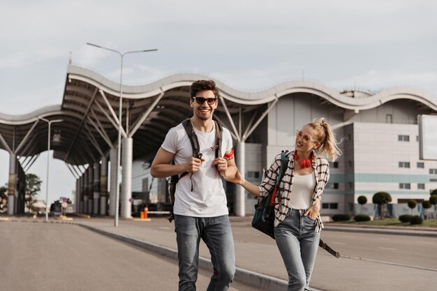 Homme brune en tee-shirt blanc et fille en chemise à carreaux sourit et marche avec des sacs à dos le long de la route Les voyageurs en lunettes de soleil parlent près de l'aéroport