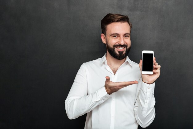 Homme brunette positif montrant smartphone sur appareil photo démontrant ou gadget publicitaire sur l'espace de copie de graphite