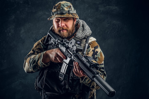 Homme brutal dans l'uniforme militaire camouflé tenant un fusil d'assaut. Photo de studio contre un mur texturé sombre