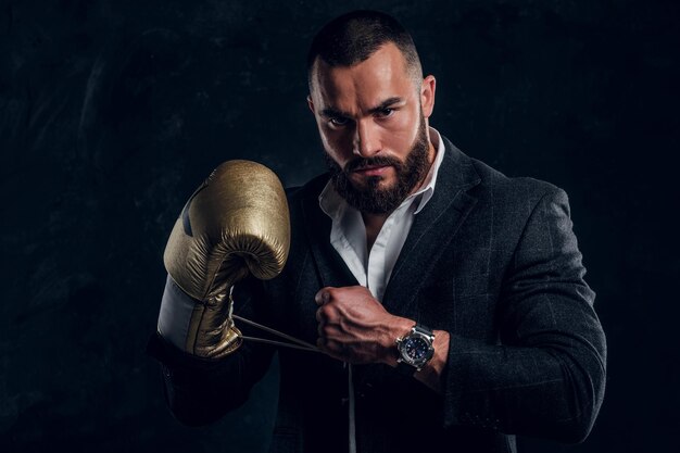 Un homme brutal et sérieux en costume et gant de boxe doré pose pour le photographe dans un studio photo sombre.