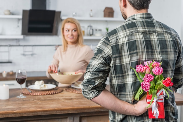 Homme avec cadeau et fleurs de dos et femme en cuisine