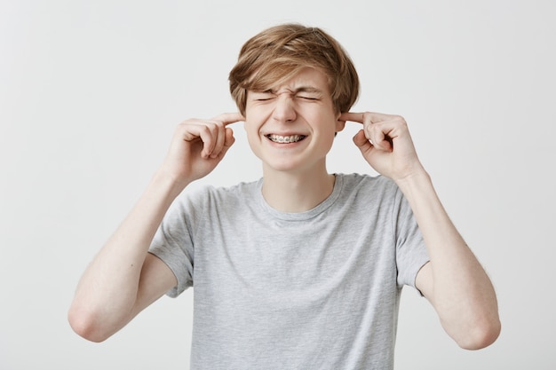 Homme caucasien agacé en colère avec des cheveux blonds serre les dents vêtu d'un t-shirt gris clair bouchant les oreilles avec les doigts, irrité par un bruit fort. Émotions, sentiments et réactions humains. Le langage du corps