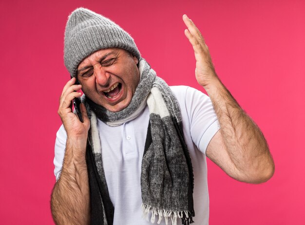 homme caucasien malade adulte en colère avec une écharpe autour du cou portant un chapeau d'hiver criant à quelqu'un au téléphone isolé sur un mur rose avec espace de copie