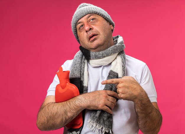homme caucasien malade adulte confiant avec une écharpe autour du cou portant un chapeau d'hiver tenant et pointant une bouteille d'eau chaude regardant de côté isolé sur un mur rose avec espace de copie
