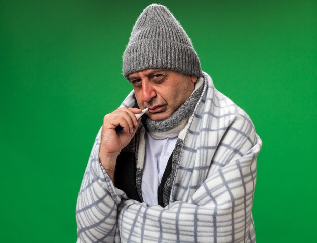 homme caucasien malade adulte désemparé avec une écharpe autour du cou portant un chapeau d'hiver enveloppé dans un plaid tenant un thermomètre dans sa bouche isolé sur un mur vert avec espace de copie