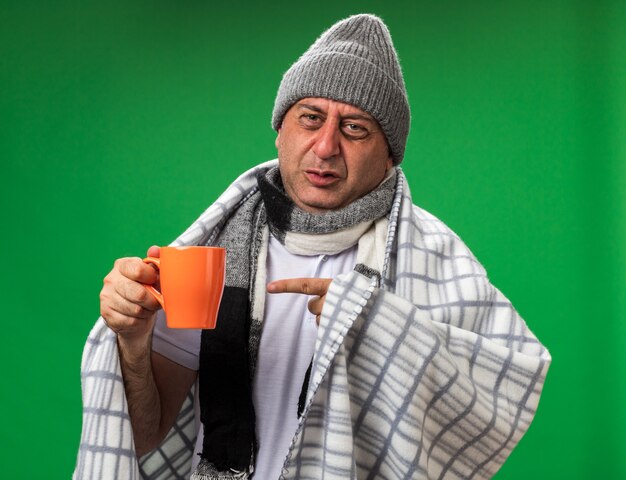 Photo gratuite homme caucasien malade adulte mécontent avec une écharpe autour du cou portant un chapeau d'hiver enveloppé dans un plaid pointant sur une tasse isolée sur un mur vert avec un espace de copie