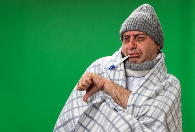 homme caucasien malade adulte mécontent avec une écharpe autour du cou portant un chapeau d'hiver enveloppé dans un plaid les pouces vers le bas tenant un thermomètre dans sa bouche isolé sur un mur vert avec espace de copie