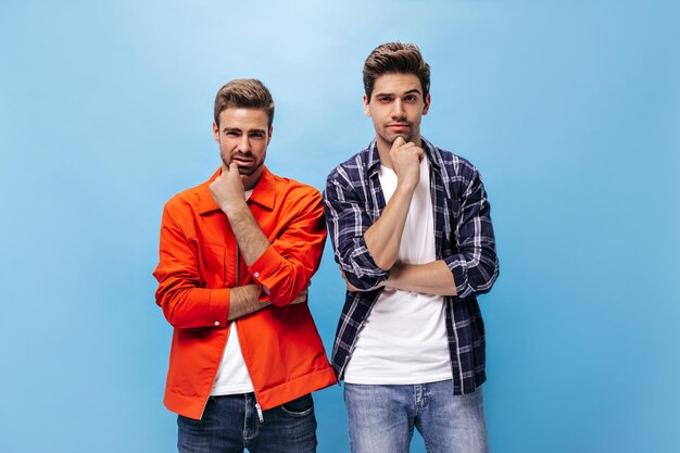 Homme charmant en jeans pose sur fond bleu Homme pensif en veste orange et mec en chemise à carreaux regarde dans la caméra sur fond bleu