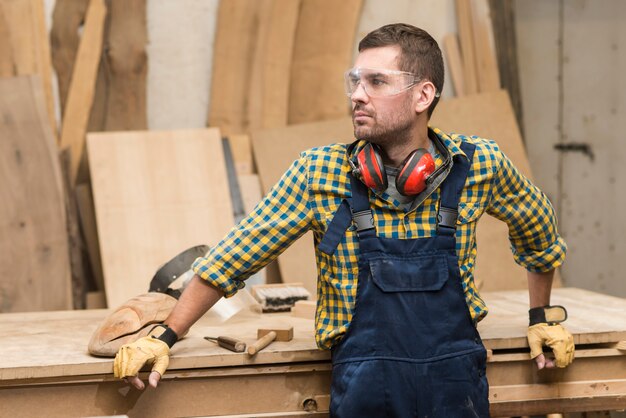 Homme charpentier avec lunettes de sécurité et protège-oreilles autour du cou, debout devant un établi en bois