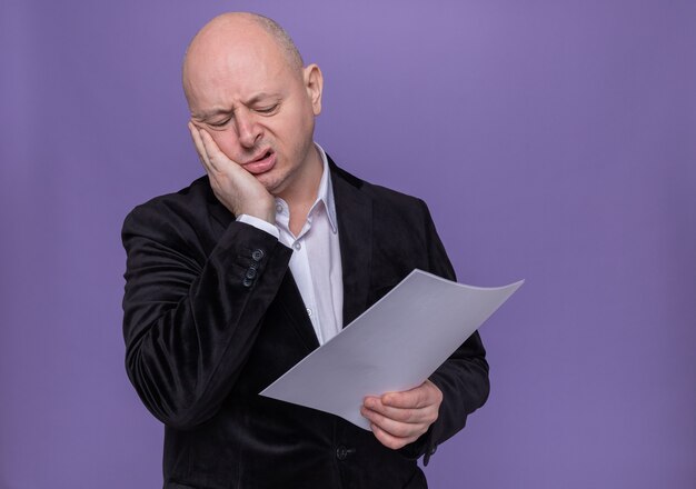 Homme chauve d'âge moyen en costume tenant une page blanche à la fatigue et s'ennuie debout sur le mur violet