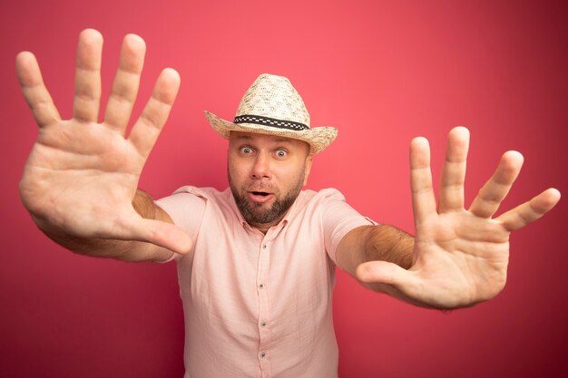 Homme chauve d'âge moyen effrayé portant un t-shirt rose et un chapeau levant les mains isolé sur rose