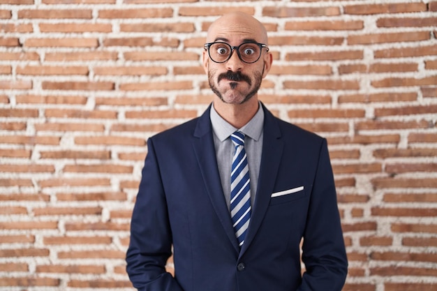 Homme chauve avec barbe portant des vêtements d'affaires et des lunettes gonflant les joues avec un drôle de visage. bouche gonflée d'air, expression folle.