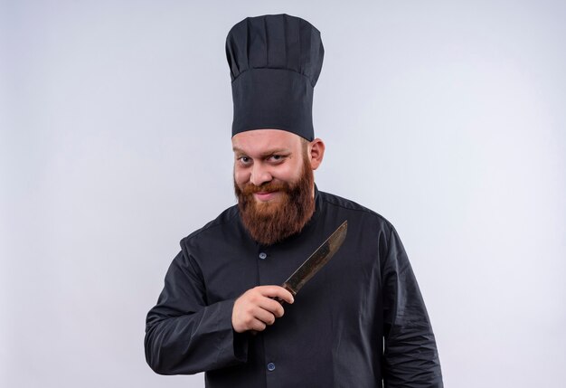 Un homme chef barbu positif et drôle en uniforme noir tenant un couteau tout en regardant la caméra sur un mur blanc
