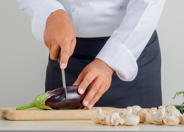 Homme chef coupe aubergine sur planche de bois en uniforme et tablier dans la cuisine