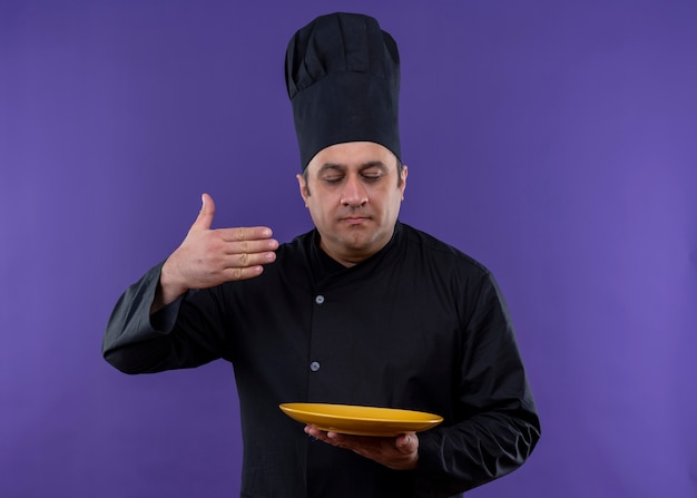 Homme chef cuisinier vêtu d'un uniforme noir et chapeau de cuisinier tenant une poêle à frire sentant une odeur agréable de plats savoureux debout sur fond violet