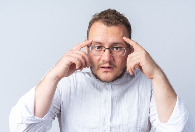 Homme en chemise blanche portant des lunettes confus et très anxieux pointant ses tempes avec les doigts debout sur un mur blanc