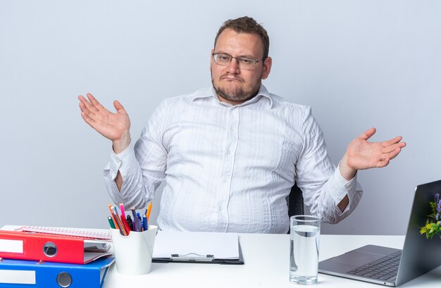 Homme en chemise blanche portant des lunettes regardant l'avant confus écartant les bras sur les côtés assis à la table avec des dossiers de bureau pour ordinateur portable et un presse-papiers sur un mur blanc travaillant au bureau