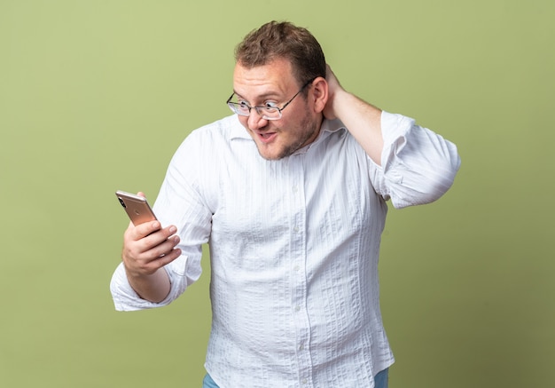 Homme En Chemise Blanche Portant Des Lunettes Tenant Un Smartphone En Le Regardant étonné Et Surpris Debout Sur Le Vert