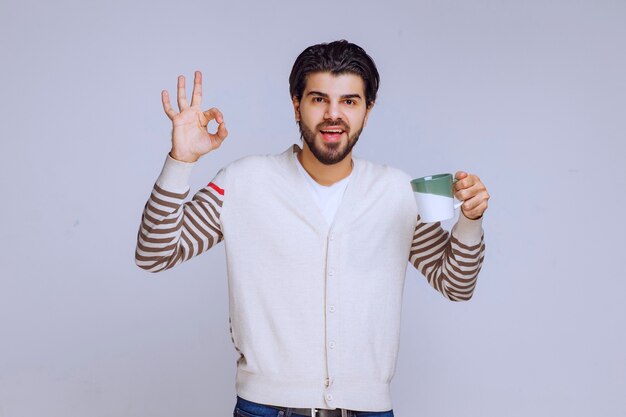 Homme en chemise blanche tenant une tasse de café et faisant un bon signe de la main.