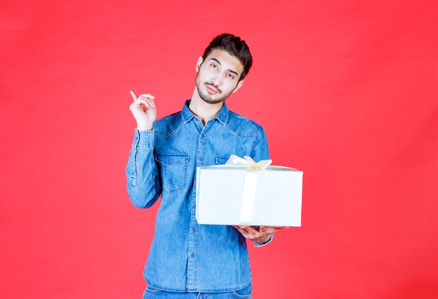 Homme en chemise en jean tenant une boîte-cadeau en argent et pointant quelque part.