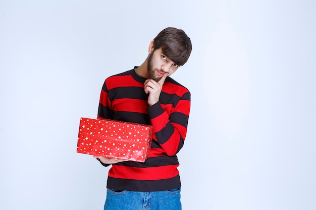 L'homme en chemise rayée rouge avec une boîte-cadeau rouge a l'air confus et réfléchi.