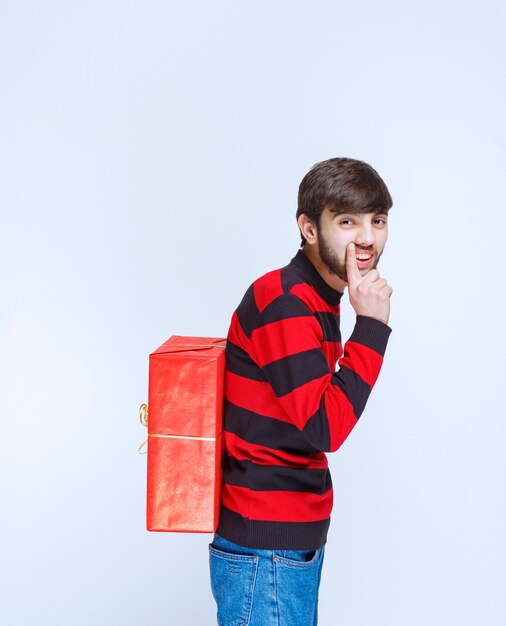 Homme en chemise rayée rouge cachant une boîte cadeau rouge derrière lui.