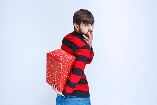 Homme en chemise rayée rouge cachant une boîte cadeau rouge derrière lui.