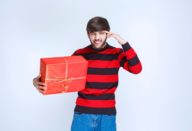 Homme en chemise rayée rouge tenant une boîte-cadeau rouge et a l'air confus et réfléchi.