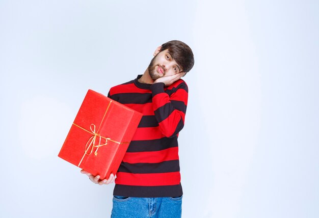 L'homme en chemise rayée rouge tenant une boîte-cadeau rouge a l'air fatigué et endormi.