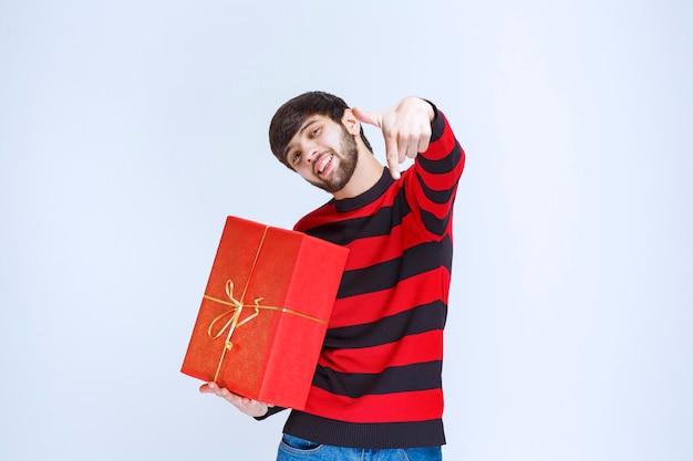 Homme en chemise rayée rouge tenant une boîte-cadeau rouge et appelant la personne juste à côté de lui.