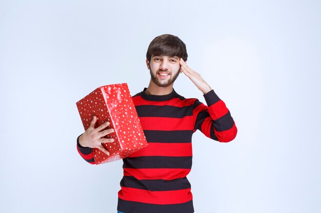 Homme en chemise rayée rouge tenant une boîte-cadeau rouge et appelant quelqu'un pour la livrer.