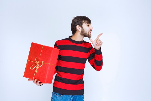 Homme en chemise rayée rouge tenant une boîte-cadeau rouge et en faisant la promotion.