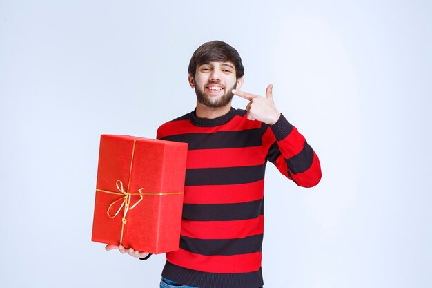 Homme en chemise rayée rouge tenant une boîte-cadeau rouge et en faisant la promotion.