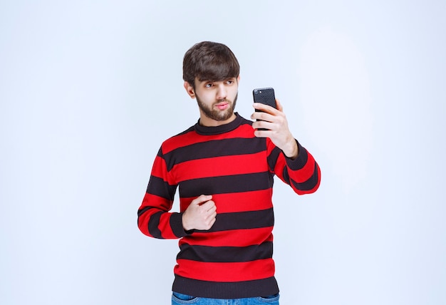 Homme en chemise rayée rouge tenant un smartphone noir et montrant qu'il apprécie les nouvelles fonctionnalités.