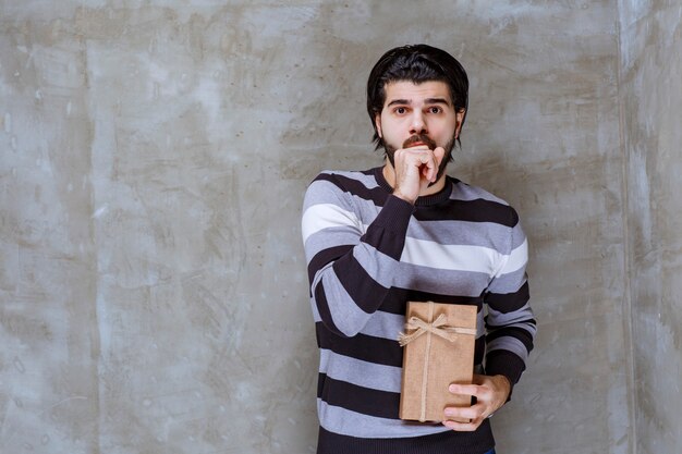 Homme en chemise rayée tenant une boîte-cadeau en carton et a l'air confus et terrifié