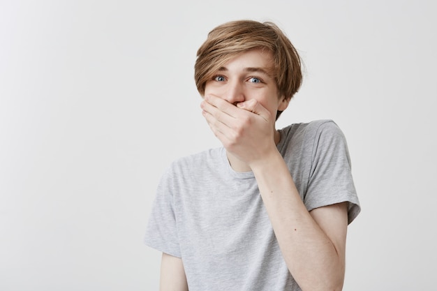 Un homme choqué, perplexe et frustré, aux cheveux blonds et aux yeux bleus, garde la main sur la bouche, surpris de faire une grosse erreur ou d'échouer à l'examen. Terrifié étonné jeune homme effrayé dans l'étonnement et la confusion