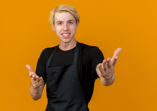 Homme de coiffeur professionnel en tablier à l'avant avec les bras comme demander et souriant debout sur un mur orange