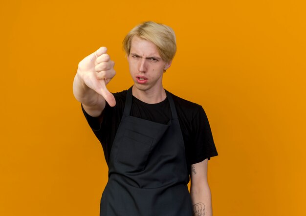 Homme de coiffeur professionnel en tablier à l'avant d'être mécontent montrant les pouces vers le bas debout sur un mur orange