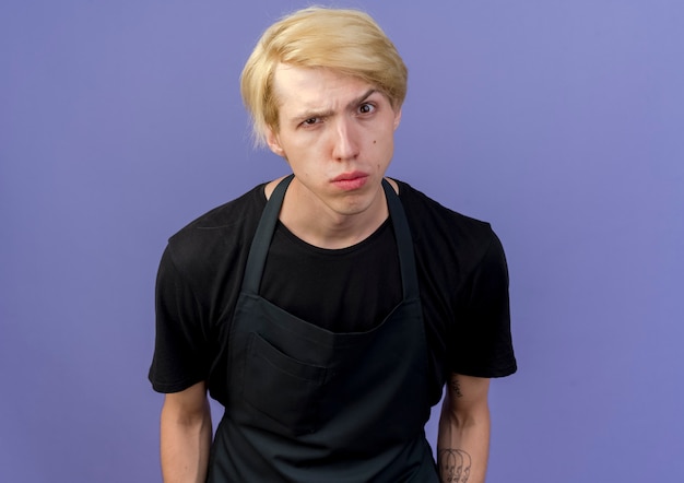 Photo gratuite homme de coiffeur professionnel en tablier à l'avant avec une expression sceptique debout sur un mur bleu