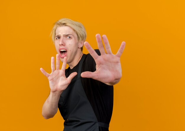 Homme de coiffeur professionnel en tablier à l'avant faisant la défense ou arrêter le geste avec les mains ayant peur debout sur le mur orange