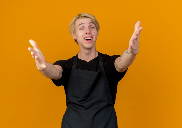 Homme de coiffeur professionnel en tablier à l'avant faisant un geste de bienvenue ouverture de larges mains souriant debout sur un mur orange