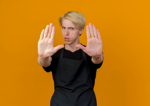 Homme de coiffeur professionnel en tablier à l'avant avec un visage sérieux faisant arrêter de chanter avec les mains ouvertes debout sur le mur orange