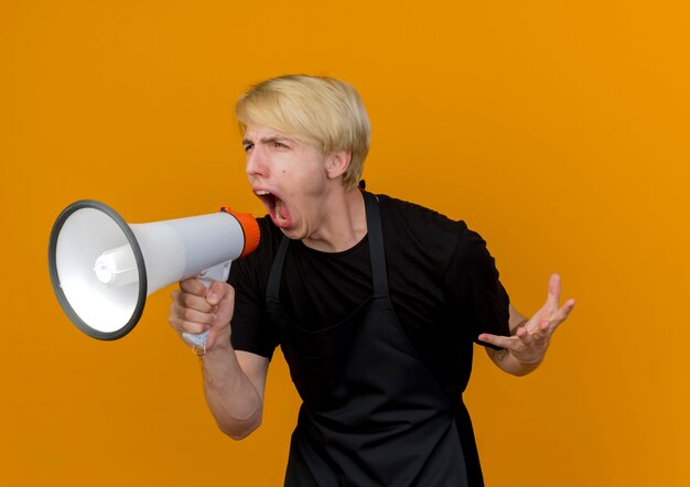 Homme de coiffeur professionnel en tablier criant au mégaphone bruyamment avec une expression agressive debout sur un mur orange