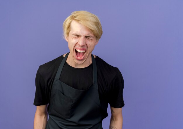 Homme de coiffeur professionnel en tablier criant avec une expression agacée debout sur un mur bleu