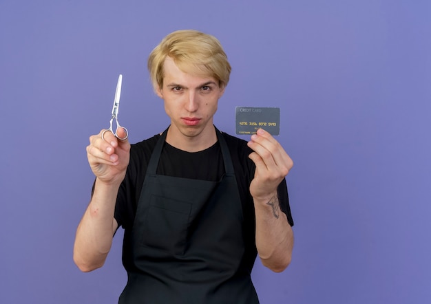 Homme de coiffeur professionnel en tablier montrant une carte de crédit et des ciseaux avec un visage sérieux