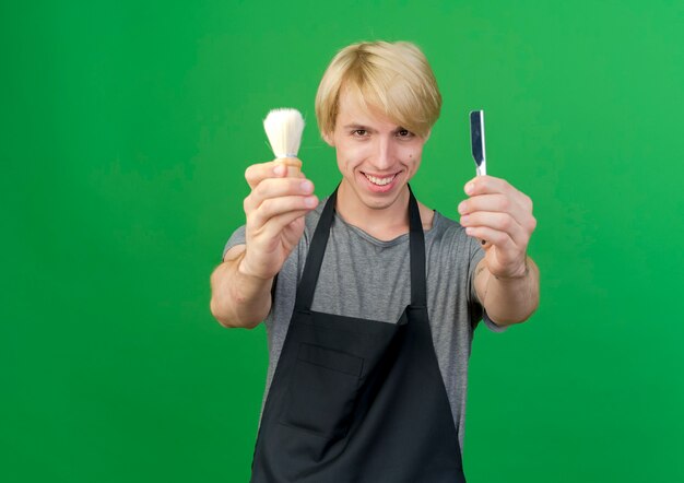 Homme de coiffeur professionnel en tablier montrant le rasoir et le blaireau souriant joyeusement