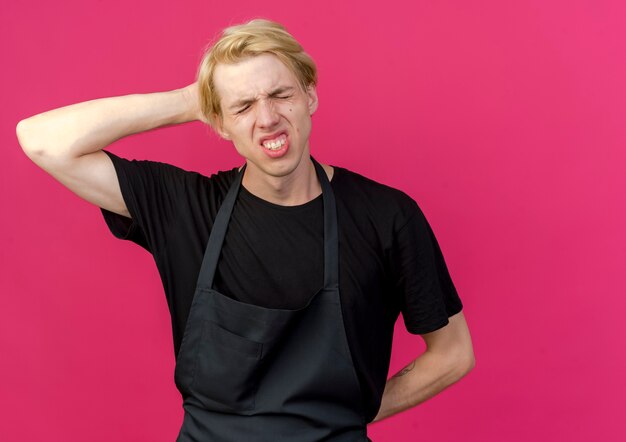 Homme de coiffeur professionnel en tablier à la recherche de mal à toucher sa tête souffrant de douleur