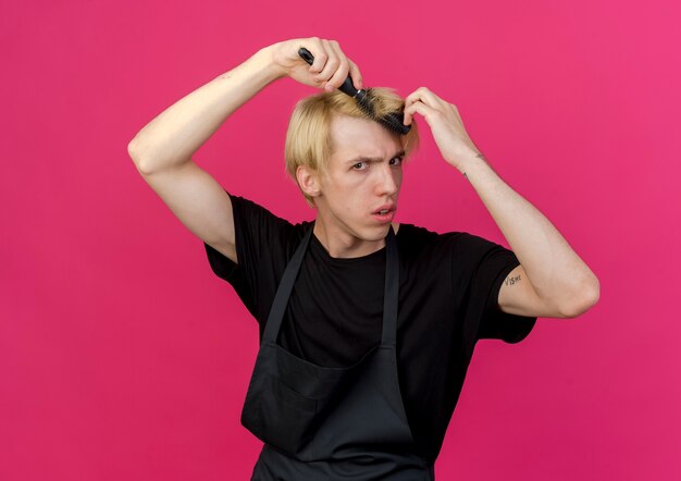 Homme de coiffeur professionnel en tablier tenant une brosse à cheveux peignant ses cheveux avec un visage sérieux